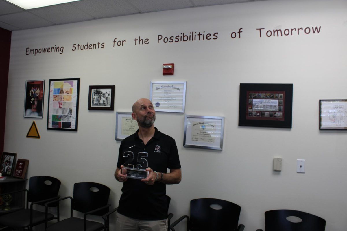 Mr. Bayer holds an award in his office. Writing on the wall reads the school motto: "Empowering Students for the Possibilities of Tomorrow."