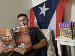 Senior Micah Lopez in his house sitting in front of a Puerto Rican flag, reading a Spanish book upside down, highlighting the confusion of the Spanish language.