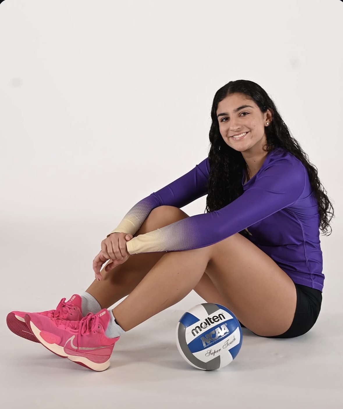 Schnell poses for a sports photo on top of a volleyball. She is dressed in her iconic purple jersey, smiling at the camera.