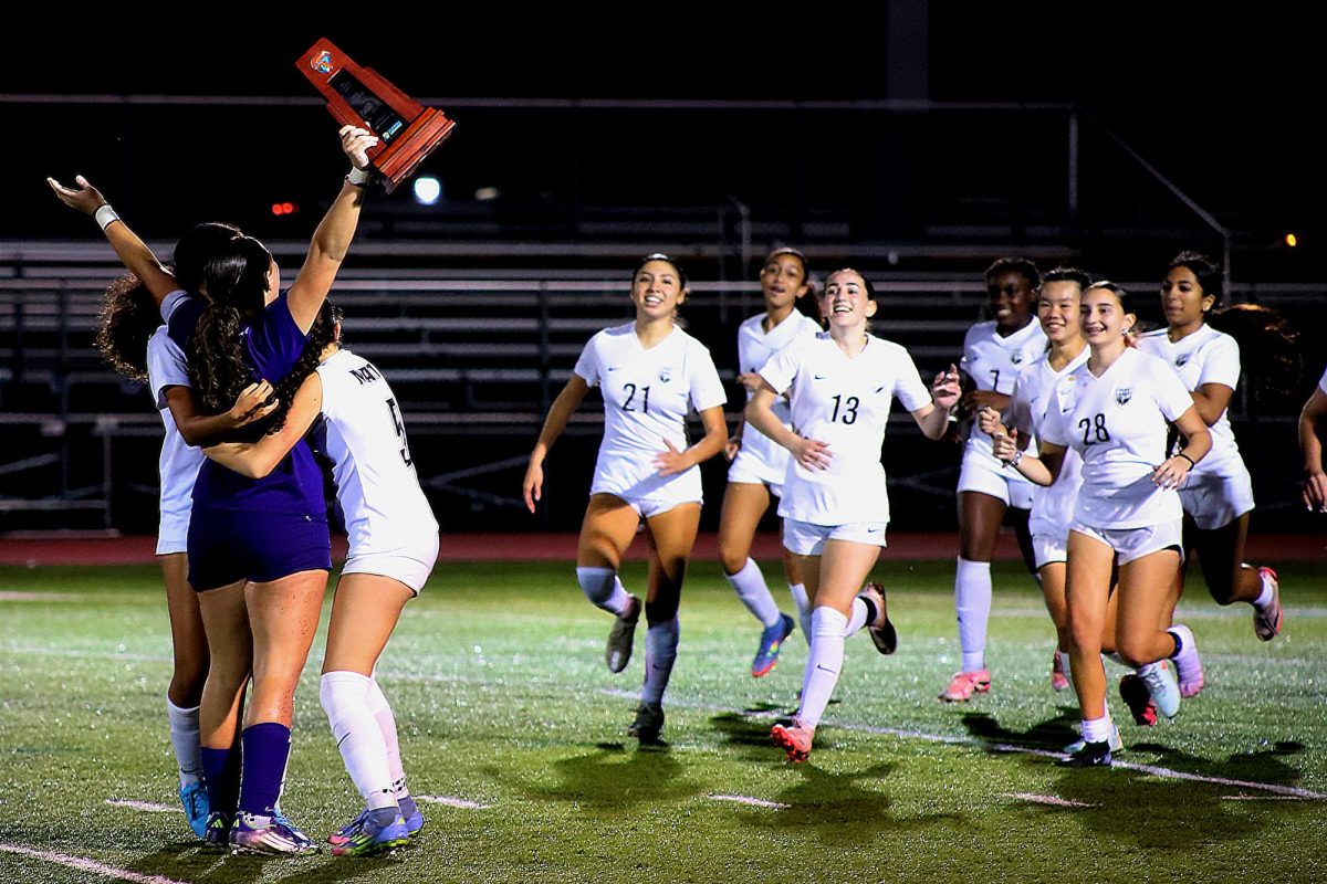 The Lady Jags captains holding the district champion trophy while their teammates run to them to celebrate.
