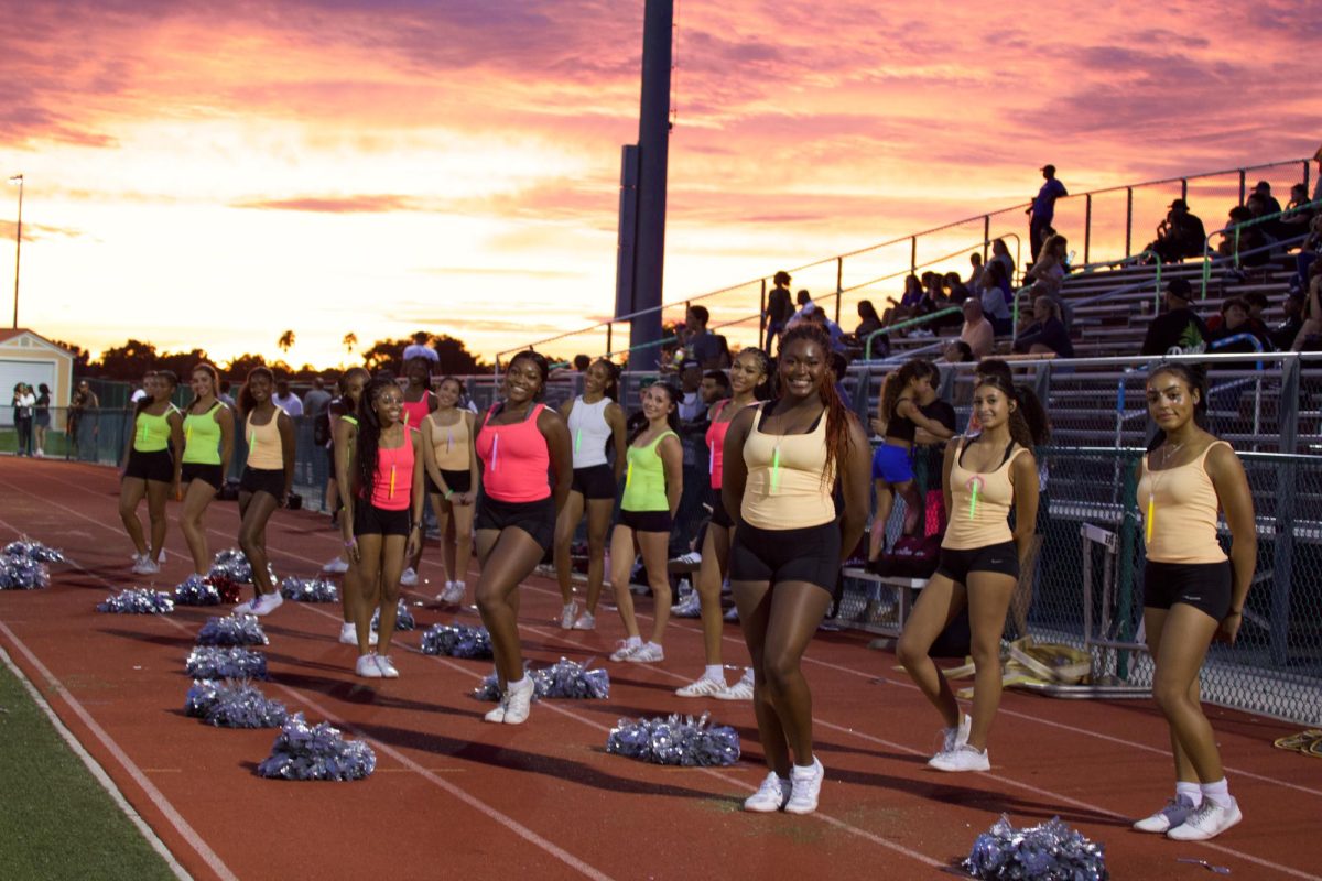 The Lady Jags cheer team standing on the field at the varsity football game as the sun sets with pink and orange clouds behind them.