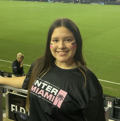 Victoria Regueiro stands in front of a soccer field and smiles at the camera as she shows off the soccer team's signature color on her face.