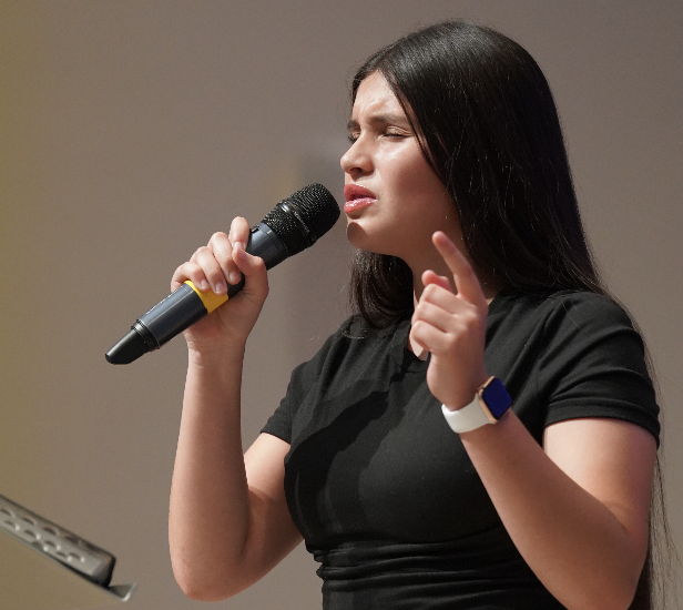 Allie Martinez sings into a microphone. Her long hair, which passes her waist, highlights her faith and worship.