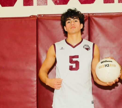 Senior Xavier Soto (#5) posing for his senior photo holding his volleyball while standing in the Jaguars home gym with his jersey on.